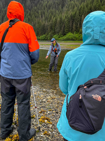 Woman guide in purple coat & gray pants stands in shallow river & lectures to nearby travelers standing in coats & rain pants on a Lindblad Alaska cruise.