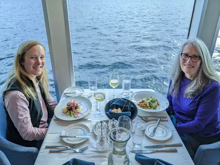2 women sit at white tablecloth dining table with colorful plated meals & water view on Nat Geo Venture small ship.