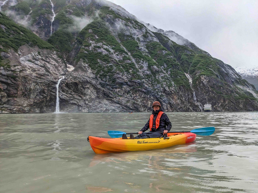 Woman in black jacket & brown hat sits in yellow, orange & red kayak with blue paddle in calm water by waterfalls & small ship.