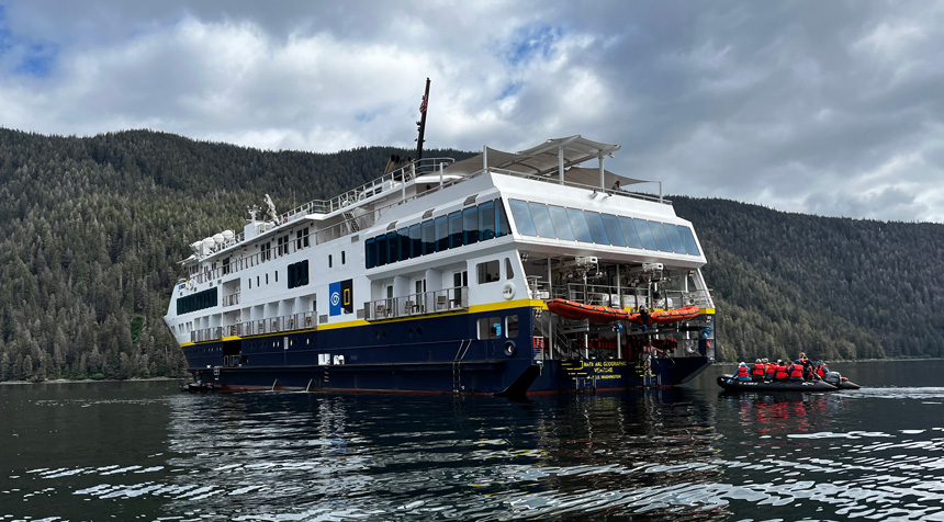 Port side & rear of Nat Geo Venture small ship as a Zodiac pulls up in calm water on a cloudy day in Alaska.