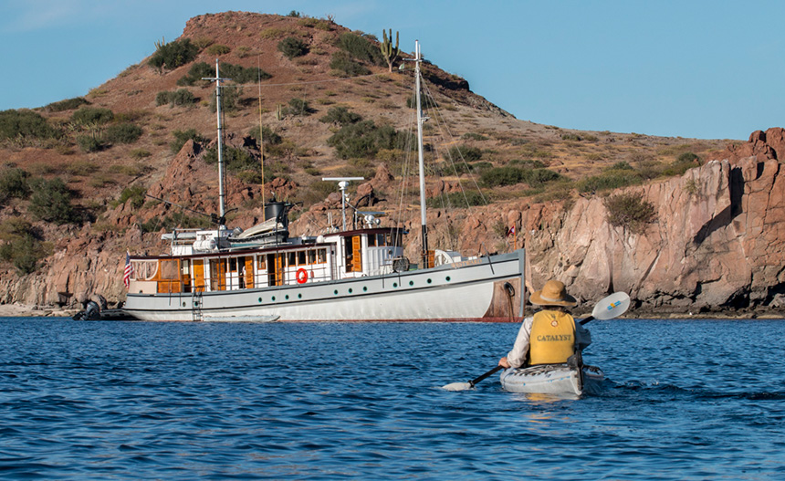 A kayaker on blue water paddling towards a historic expedition cruise yacht with rocky cliffs in the background. 