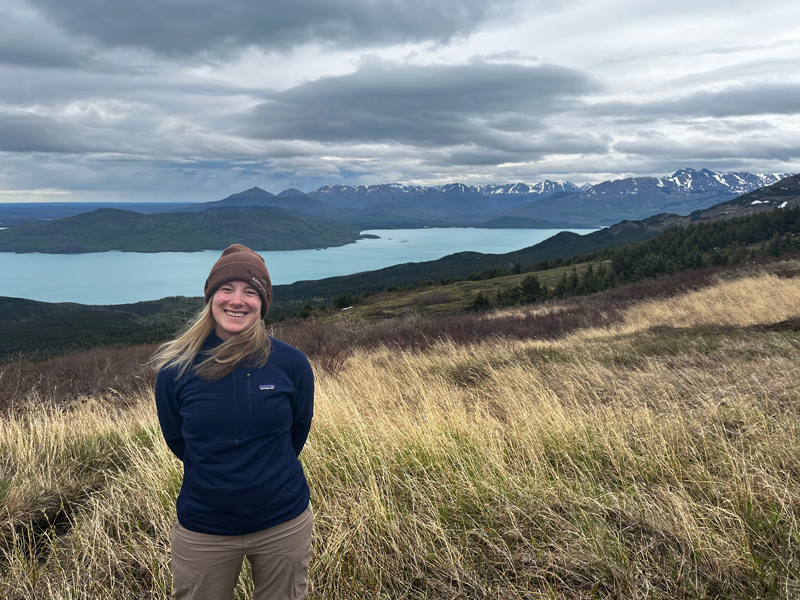 Woman in tan pants, dark blue sweater & brown knit hat stands on grassy mountaintop overlooking turquoise lake in Alaska.
