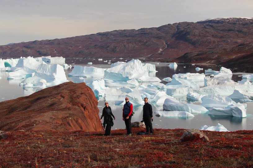 In East Greenland guests stand on bright red hillside overlooking a bay  in Scoresby Sund filled with massive floating icebergs.