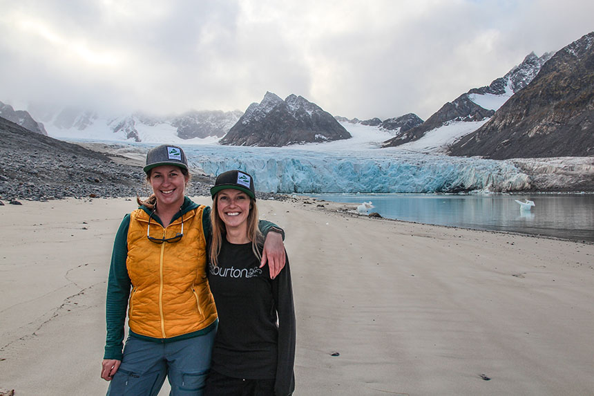 Two female travelers in AdventureSmith branded hats stand arm over shoulder of another on a beach in front of a glacier