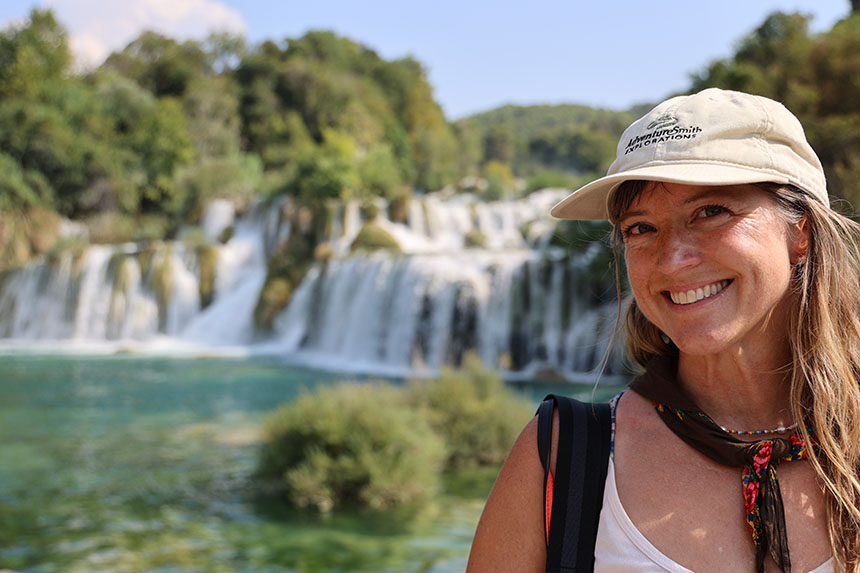 A woman in an AdventureSmith Explorations tan hat smiles standing in front of the waterfalls of Krka National Park in Croatia