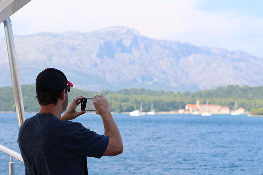 A man on a small cruise ship deck seen from behind in a dark t-shirt photographs a mountainous coastline passing by his ship