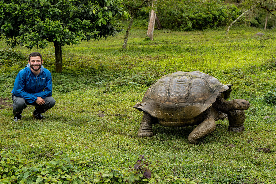 A male traveler in a blue jacket crouching in a grass covered field next to a giant Galapagos tortoise.