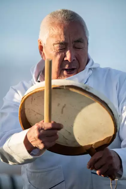 Man from remote Greenland settlement of Ittoqqortoormiit dressed in all white traditional Inuit clothing sings and plays instrument. 