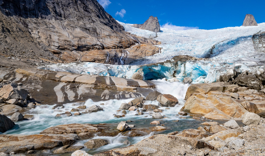 A sweeping wide glacier flows to the mouth of the rushing water flanked by tan rocks and mountains during a South Greenland cruise.
