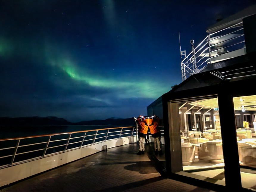 Guests stand on deck of ship staring up at the green strip of lights across the  dark night sky, it the aurora borealis in Greenland.  
