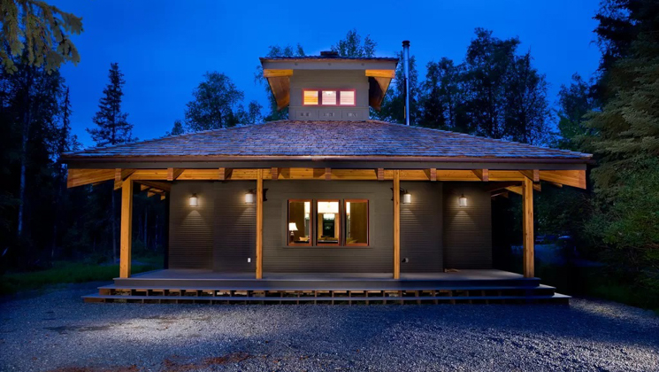 Small 1-level cottage at dusk with covered porch, 1 large window & exterior accent lights, surrounded by forest in Alaska.
