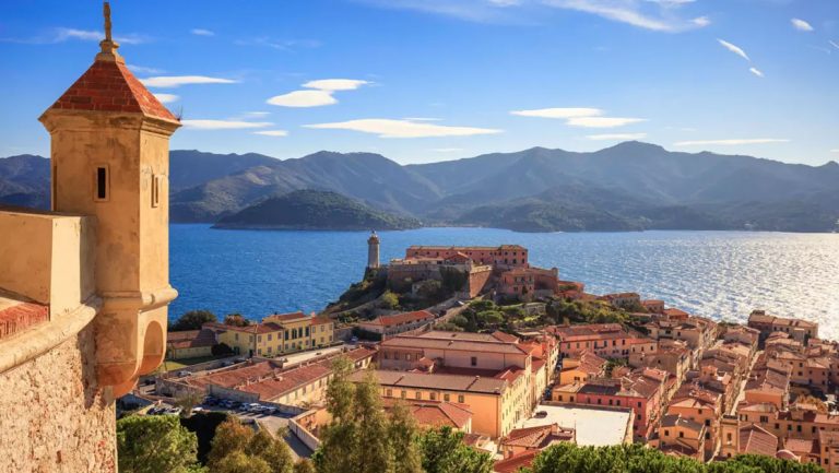 Tan stone tower sits on corner of building overlooking seaside town with terracotta roofs & rolling green hills across the water.