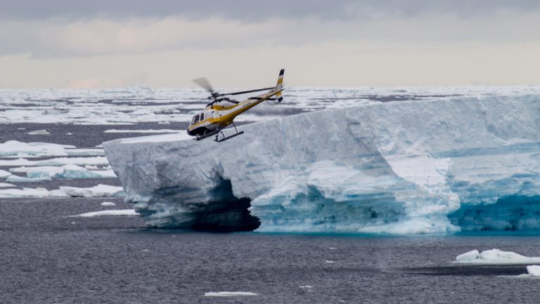 Black, white & yellow helicopter flies above large blue shelf iceberg on the Remote Weddell Explorer Antarctic cruise.