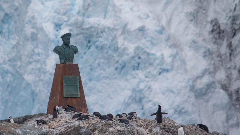 Bust of Sir Ernest Shackleton sits atop rusty platform with penguins underneath & large iceberg behind at Point Wild.