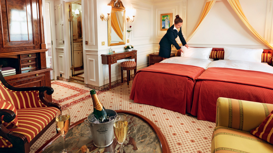Category 6 cabin on Sea Cloud II: Lindblad with double bed being made up by female steward, red & gold accents & champagne.