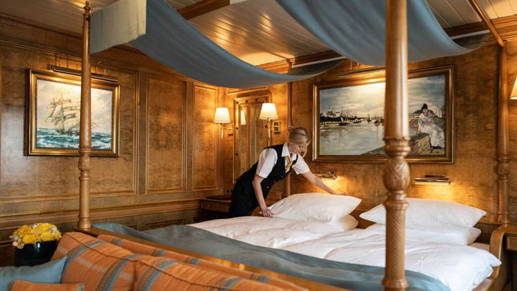 Female steward makes up 4-poster bed in Category 7 cabin with wood walls & peach couch on Sea Cloud II: Lindblad sailing ship.