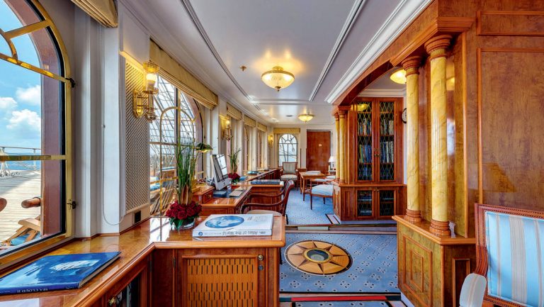 Sea Cloud II: Lindblad library with wood cabinets, & desks, large forward-facing windows & light blue carpet with compass rose.