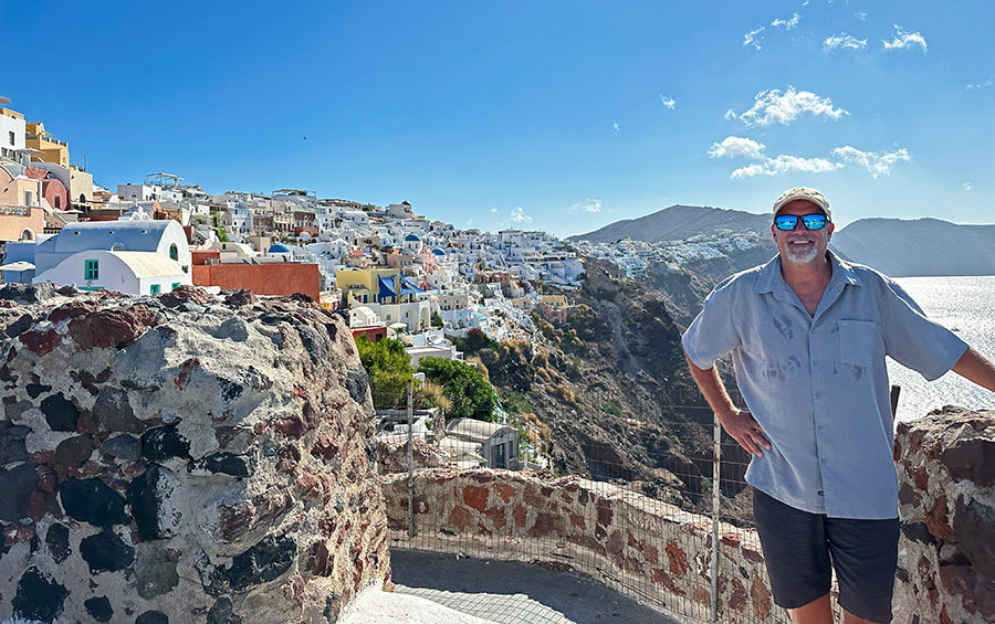 A man standing on a rocky path above a village with white houses surrounded by the Mediterranean Sea.  
