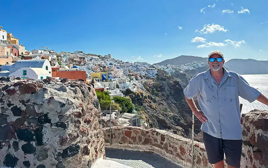 A male traveler in a blue shirt and sunglasses standing in front a Greek port village with white houses on a cliff and the Mediterranean Sea in the background. 