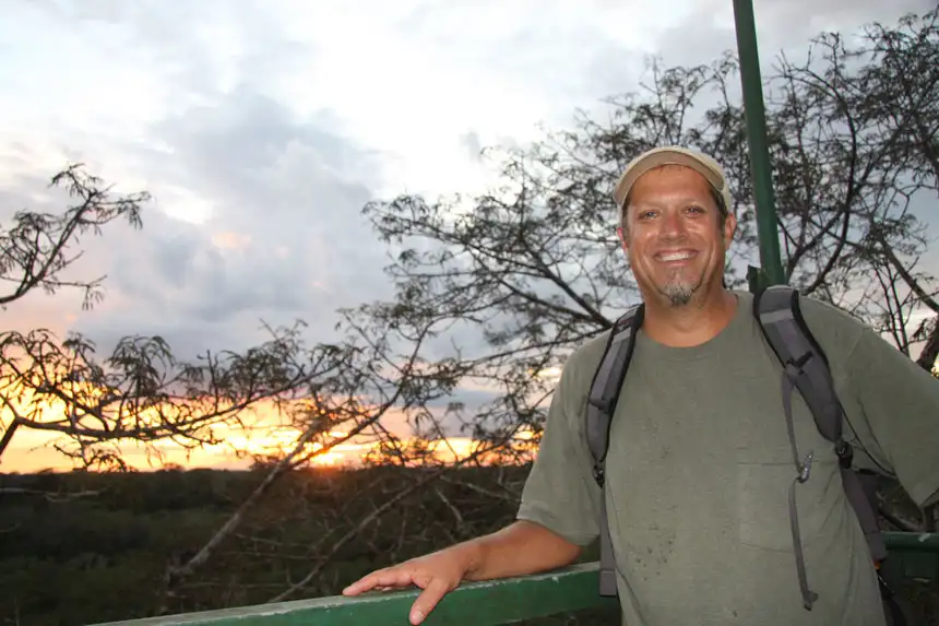 Man in tan hat & green t-shirt with gray backpack stands on a treetop rainforest tower at sunset in the Ecuadorian Amazon.