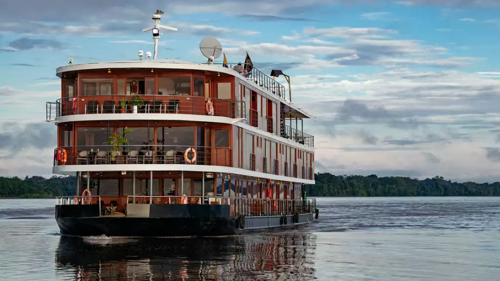 Small river cruise ship tan and wood in design with white roofs and a black bottom cruises the amazon on a calm day