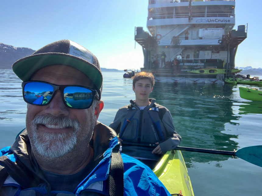 Father in sunglasses & blue jacket paddles tandem kayak with son sitting behind on a sunny day in Alaska.