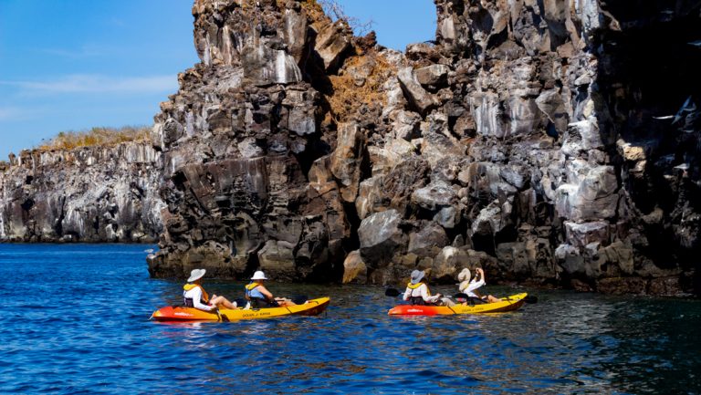 2 tandem red & yellow kayaks with duo paddlers drift by chunky bird cliffs in calm sea on a Cormorant II Galapagos cruise.