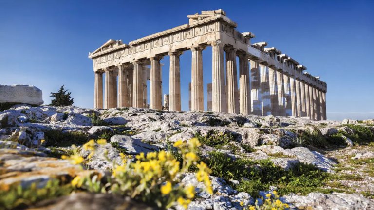 Acropolis's stone pillars create an open-air ancient building sitting atop mossy rocks and yellow flowers in the sun in Athens.