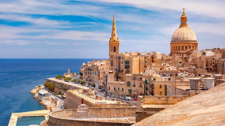 Malta Island in Italy with tan stone buildings, bell tower & dome beside turquoise sea under sunny skies.