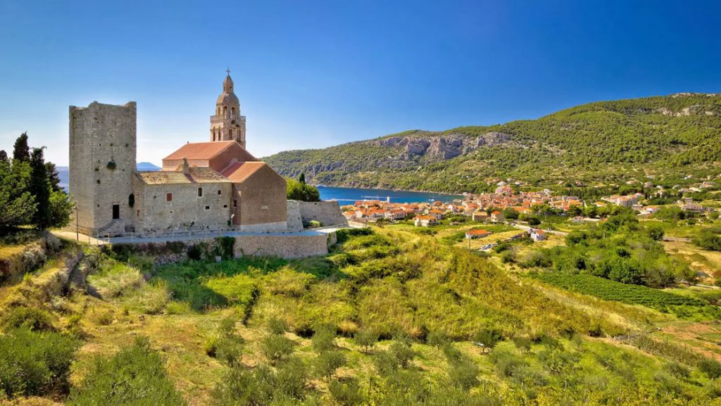 Medieval castle with tan stone & red roof sits among grassy fields by water in sun, seen on a cruise to Croatia and Italy.