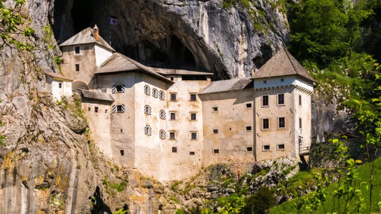 Large medieval stone building with pointed roofline sits outside cave opening among stone & green forest in Slovenia.
