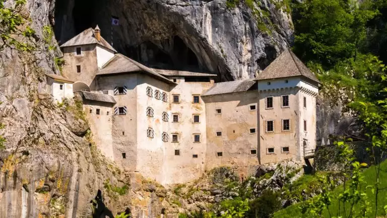 Large medieval stone building with pointed roofline sits outside cave opening among stone & green forest in Slovenia.
