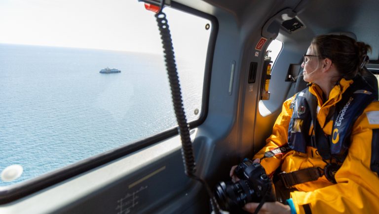 Woman in yellow jacket & PFD with camera in hands sits in helicopter looking at small expedition ship out the window & smiles.