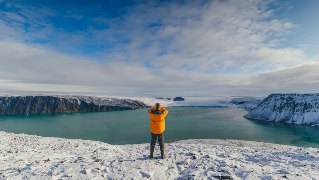 Greenland glacier cruise traveler in yellow coat stands atop snow-covered mountain looking down on turquoise fjord & large glacier.