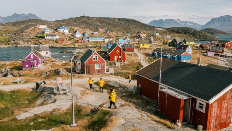 2 Greenland glacier cruise guests in yellow jackets walk through Arctic town with colorful homes on a sunny day.
