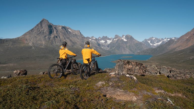 2 Greenland glacier cruise travelers in yellow coats stand over mountain bikes on top of green overlook with fjord below.