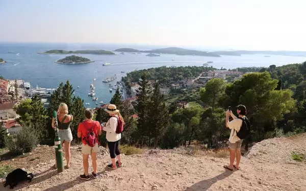 Four travelers stand on a dirt hilltop in Hvar Croatia taking pictures of the town and port below filled with boats