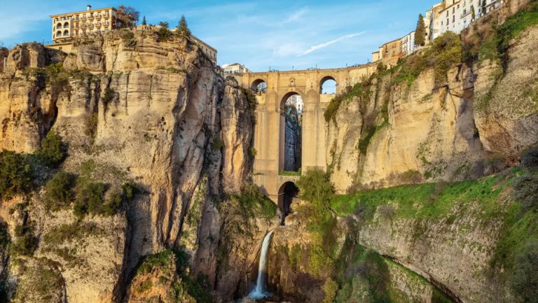 Waterfall in stone canyon sits under tall bridge with hanging moss & stone buildings in sun on an Iberian Peninsula cruise.