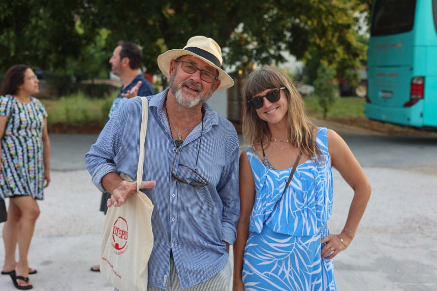 An older male travelers in a Panama hat and blue button-up shirt stands beside a younger female traveler in a blue dress with sunglasses on.