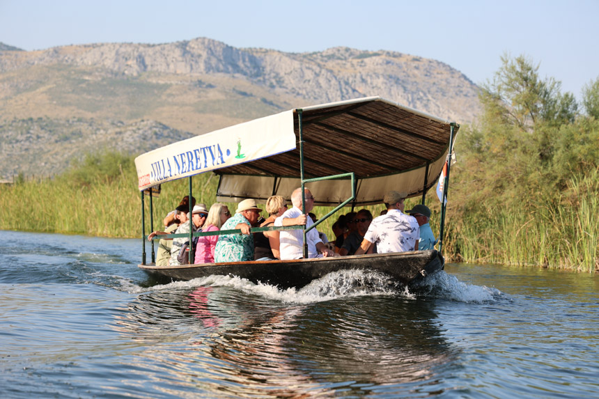 A wooden river boat with a shade top cruises towards the camera full of travelers and mountains behind it