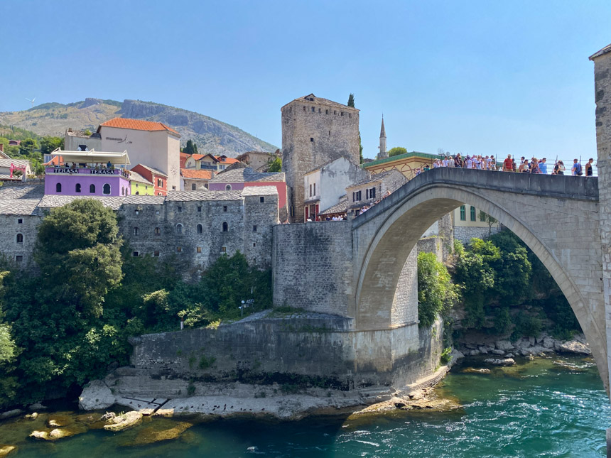 The stone Mostar bridge seen from its profile view with colorful buildings on the bank of the river and lots of people walking over the bridge