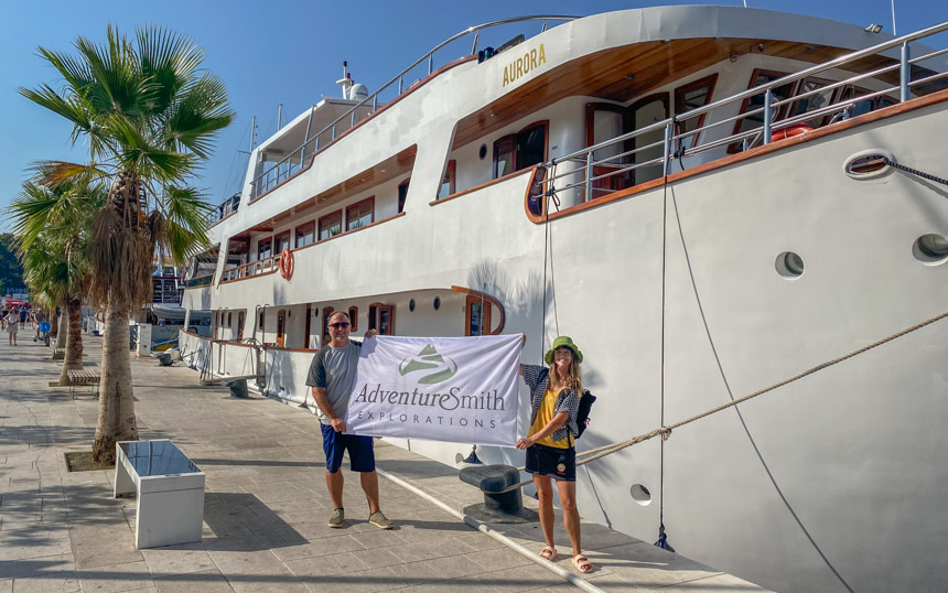 A male and female traveler stand in front of a large white yacht docked streetside in Split Croatia holding a white AdventureSmith flag between them