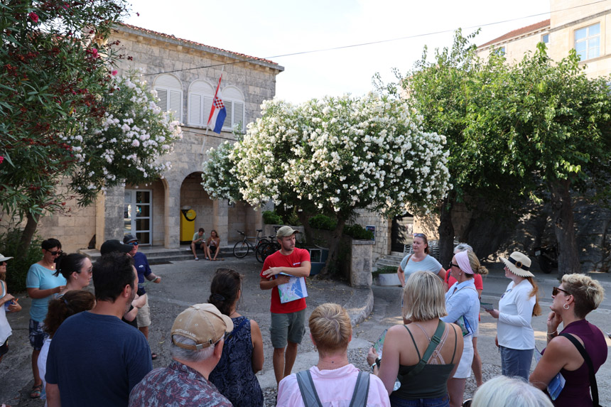 A guide in a red shirt holds a map and speaks to a group of passengers among blooming trees in Korcula Croatia