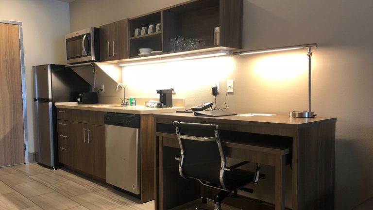 Kitchenette at Kodiak Compass Suites with stainless steel refrigerator & dishwasher, wood accent cupboards, sink & small desk.