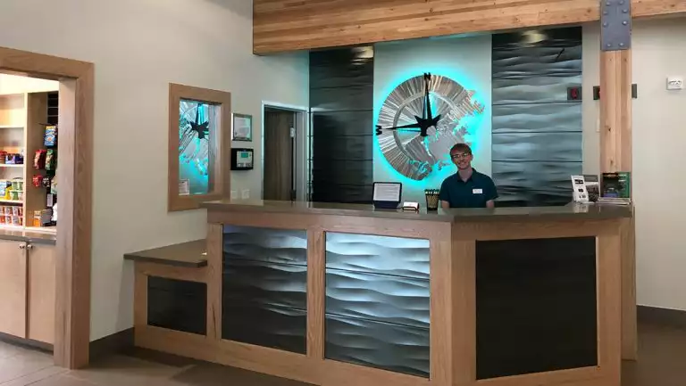 Young man stands behind reception desk at Kodiak Compass Suites that's decorated in modern wood & metal motif, beside store.