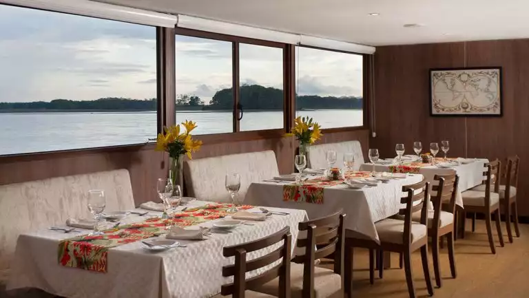 Dining room on Manatee Amazon Explorer ship with 4-top tables set for dinner in white linens, tropical runners & yellow flowers.