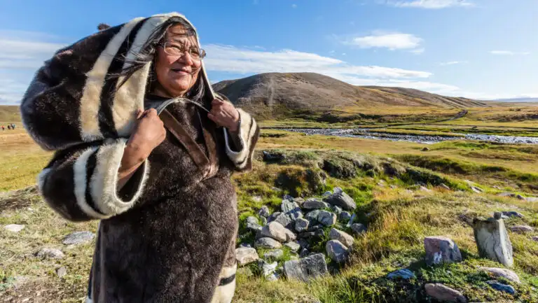 Native woman in hooded overcoat made from dark animal fur stands in green tundra backed by mountains on a sunny day.