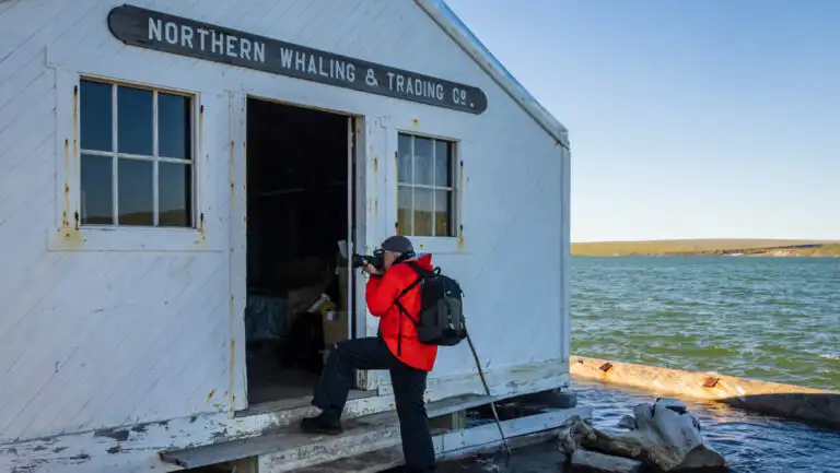 Northwest Passage traveler in red jacket stands outside door of historic whaling company building at water's edge & takes photos.