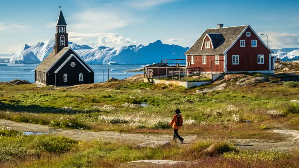 Northwest Passage traveler walks a lone road among green tundra & dark red church & home by fjord backed by snowy peaks.