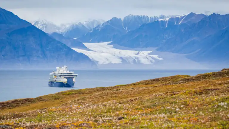 Small, modern, blue & white expedition ship sits in calm fjord beside rocky mountains, white glacier & green riverbanks.